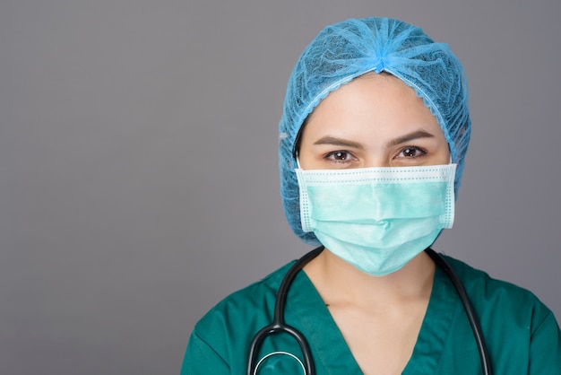 Young confident woman doctor in green scrubs is wearing surgical mask over grey 