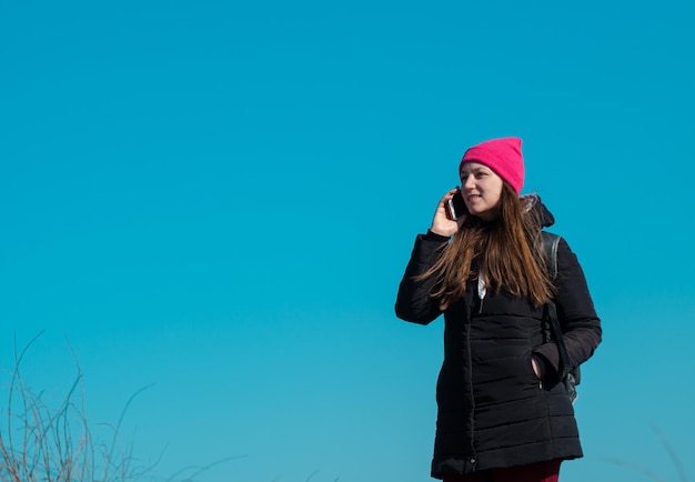 Photo young confident woman in pink hat smartphone in hand calling friend walking on blue sky background