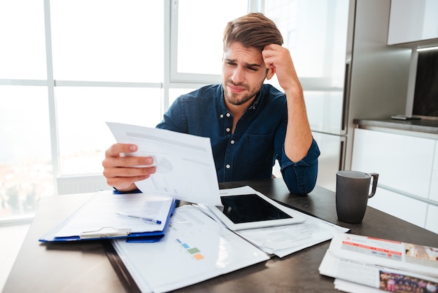 Giovane uomo confuso analizzando le finanze a casa tenendo la testa con la mano e guardando i documenti