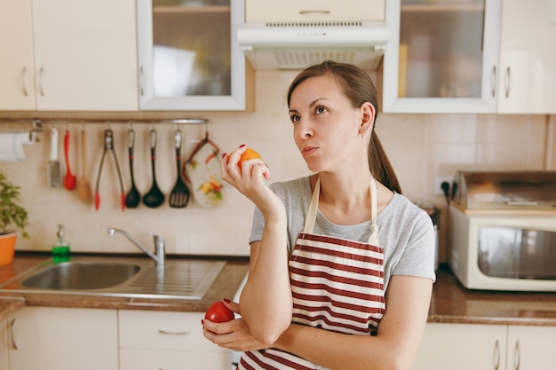 エプロンを着た若い混乱した物思いにふける女性は、台所で赤または黄色のトマトを選ぶことにしました。ダイエットの概念。健康的な生活様式。家で料理。食べ物を用意します。