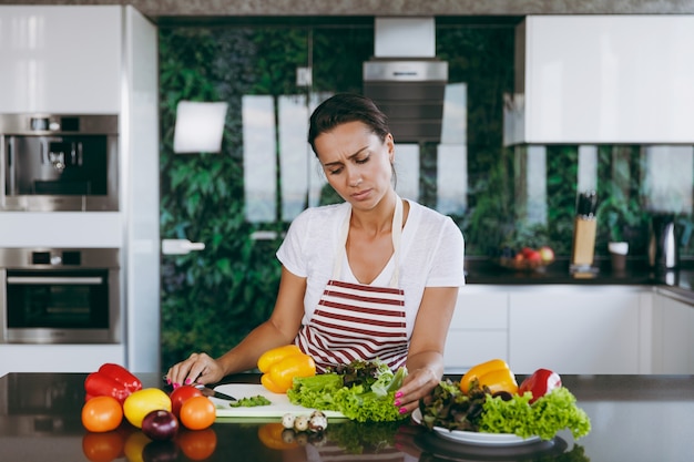 La giovane donna confusa e premurosa in grembiule decide cosa cucinare in cucina