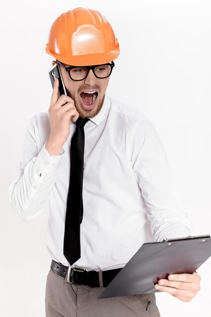 Young construction engineer in orange helmet with folder talking on phone on white background