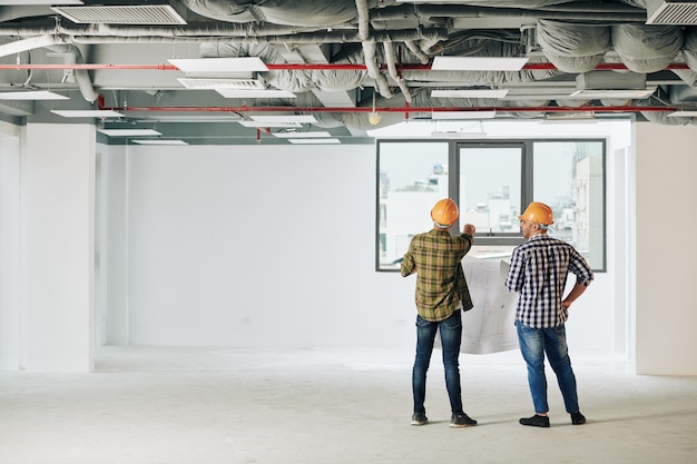 Young construction engineers in plaid shirts and hardhats holding blueprint and looking at city through window