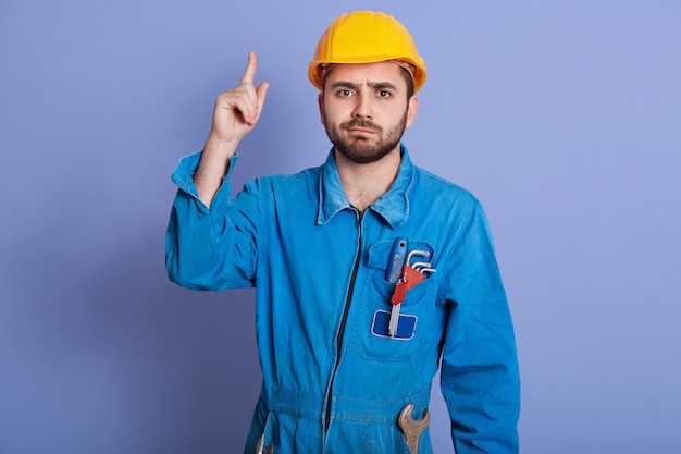 young construction worker with tools in pocket, dresses yellow helmet and uniform pointing up