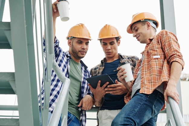 Young construction workers in hardhats drinking take away coffee and checking e-mail from contractor on tablet computer