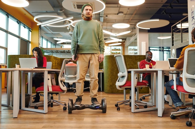 Young contemporary businessman on gyroscope moving along large open space office between desks where his colleagues using computers