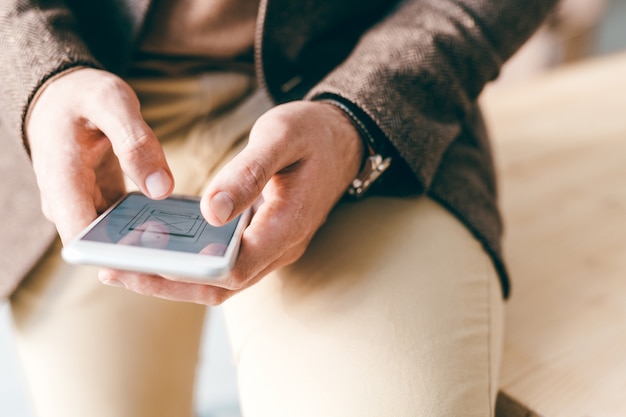 Young contemporary businessman sitting on edge of table while scrolling through electronic documents in smartphone