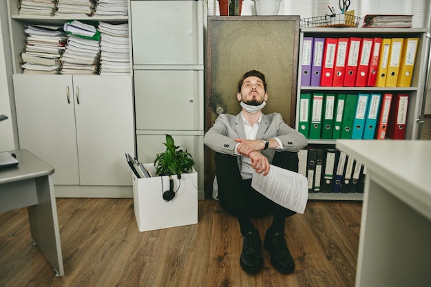 Фото Молодой современный бизнесмен с задумчивым выражением лица и бумагами в руках, сидя на полу против сейфа в офисе