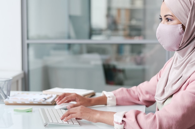 Young contemporary businesswoman in hijab and protective mask sitting by desk in front of computer and browsing in the net in office