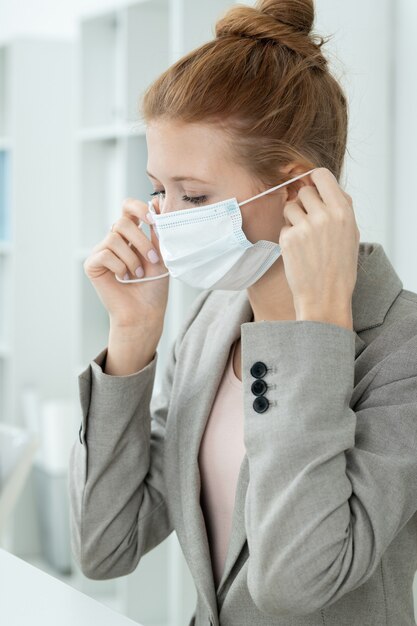 Young contemporary businesswoman putting protective mask on face to protect herself from infection during work in office environment