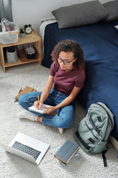 Foto giovane studente contemporaneo a gambe incrociate seduto davanti al laptop e prendendo appunti mentre ascolta l'insegnante durante la lezione online