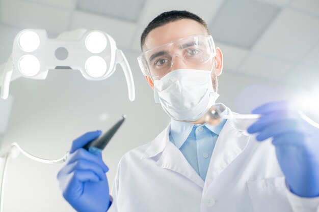 Young contemporary dentist in mask, gloves and whitecoat holding drill and mirror while bending over patient before medical procedure