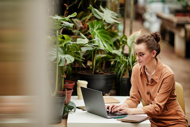 Молодая современная женщина-дизайнер или владелица малого бизнеса, работающая в Интернете, сидя за столом перед ноутбуком в цветочном магазине