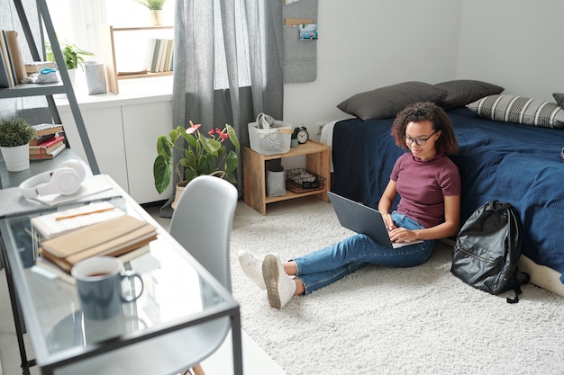 Фото Молодая современная студентка в повседневной одежде сидит на полу у кровати с ноутбуком на ногах во время онлайн-урока в домашней обстановке