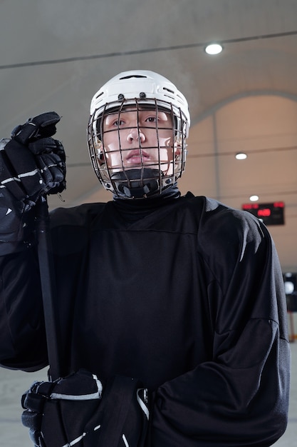 Photo young contemporary hockey player in black sports uniform, gloves and protective helmet looking forwards while standing on ice rink