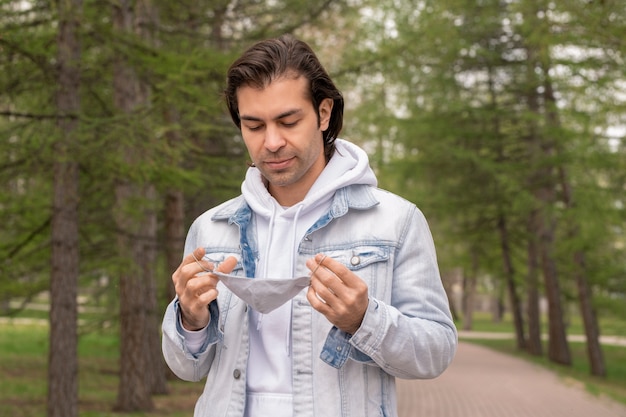 Young contemporary man in hoodie and denim jacket holding protective mask