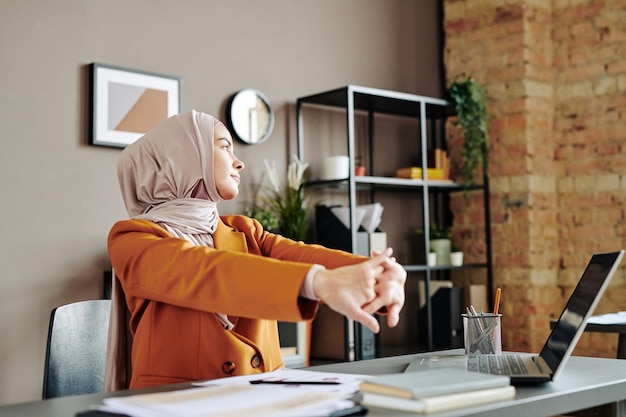 Young contemporary muslim businesswoman doing stretch exercise by workplace