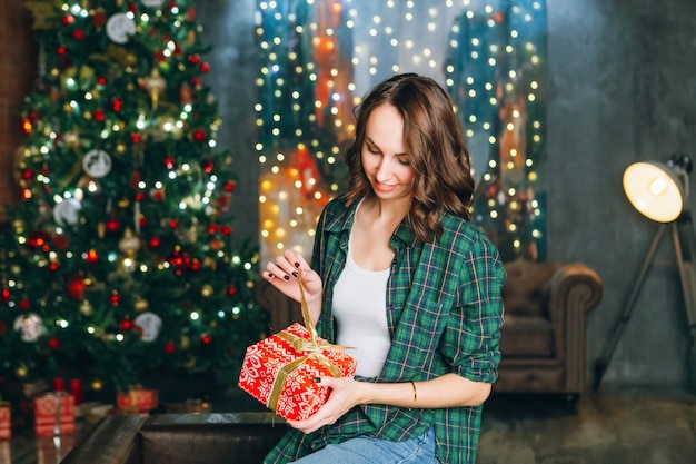 Young contented beautiful brunette woman holds a gift box in her hands at the Christmas tree