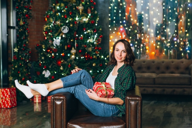 La giovane bella donna castana soddisfatta tiene un contenitore di regalo nelle sue mani all'albero di natale