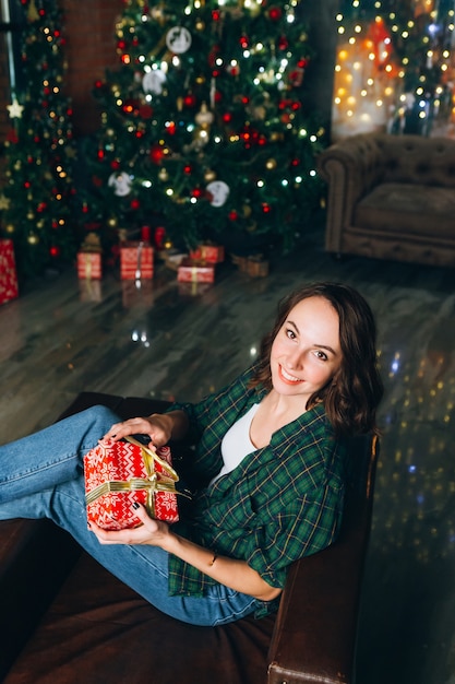 Young contented beautiful brunette woman holds a gift box in her hands at the Christmas tree