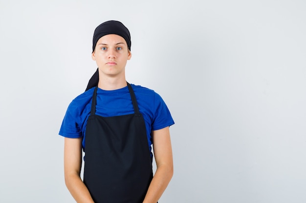 Young cook looking at front in t-shirt, apron and looking pensive. front view.