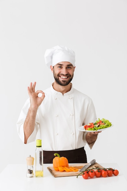 young cook man in uniform showing ok sign and holding plate with vegetable salad isolated over white wall