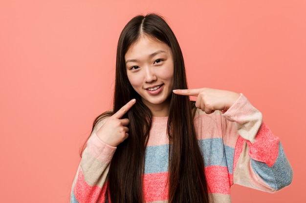 Young cool chinese woman smiles, pointing fingers at mouth.