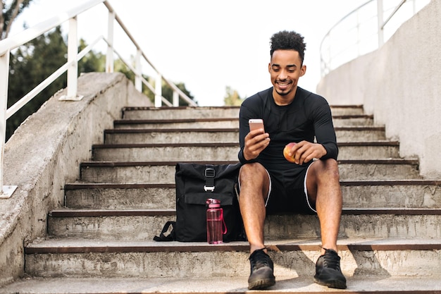 Young cool happy brunet darkskinned man in black shorts and stylish sport tshirt sits on stairs outside looks at phone screen and holds apple