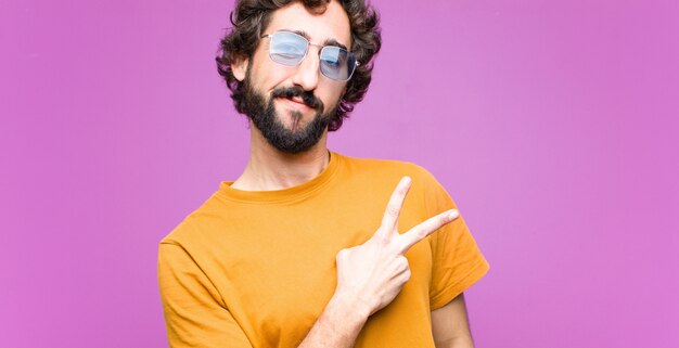 Young cool man feeling happy, positive and successful, with hand making v shape over chest, showing victory or peace against wall