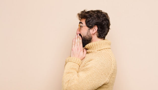 Young cool man looking happy, cheerful, lucky and surprised covering mouth with both hands against flat wall