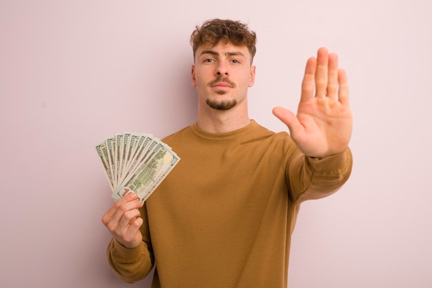 Young cool man looking serious showing open palm making stop gesture dollar banknotes concept