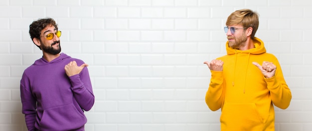 Young cool men smiling cheerfully and casually pointing to copy space on the side, feeling happy and satisfied over white tiles wall