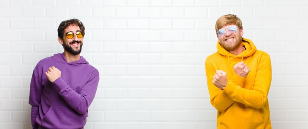 Young cool men smiling cheerfully and celebrating, with fists clenched and arms crossed, feeling happy and positive against white tiles wall