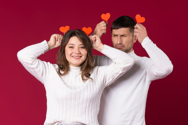 A young couple are fooling around on a red background with handmade cardboard hearts.