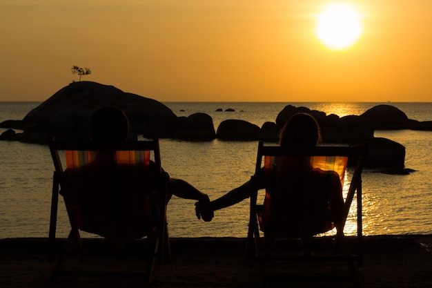 Young couple on beach hammocks holding hands while enjoying splendid sunset by the sea with rocks over water on the background.