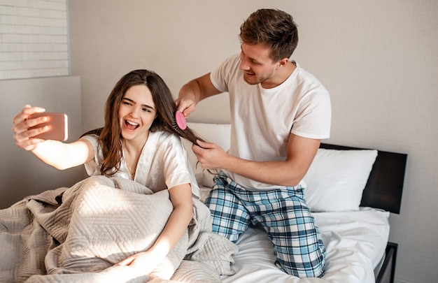 Young couple in the bed. Smiling beautiful man and woman are fooling around while making a selfie, happy morning