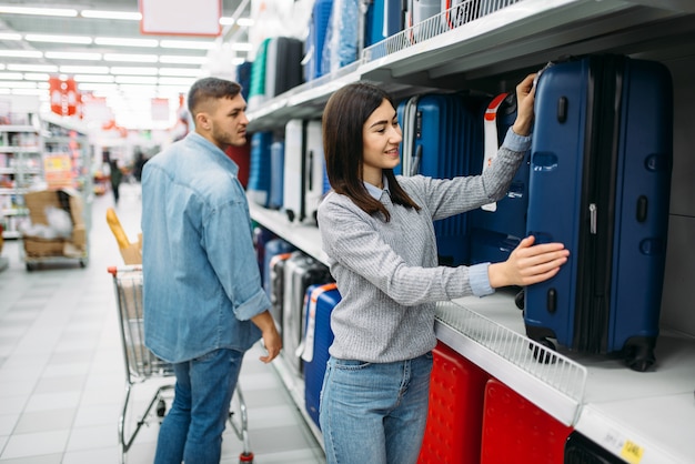 Young couple choosing suitcase in a supermarket, family shopping. Customers in shop, buyers in store looking on travel bags