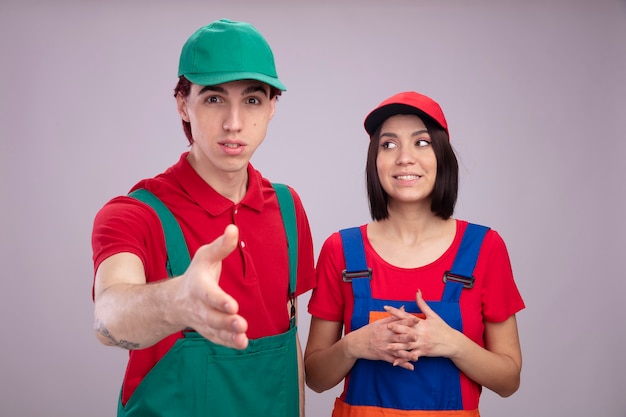 Young couple in construction worker uniform and cap impressed guy looking at camera doing greeting gesture excited girl keeping hands together looking at side isolated on white wall