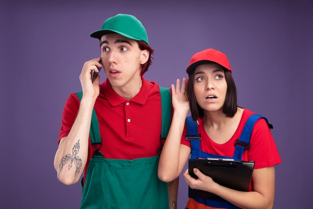 Young couple in construction worker uniform and cap impressed guy talking on phone looking at side curious girl holding clipboard looking up listening to phone conversation isolated