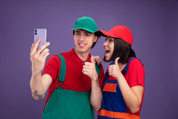 Young couple in construction worker uniform and cap taking selfie together smiling guy excited girl showing thumb up girl keeping hand on guy's shoulder isolated on purple wall