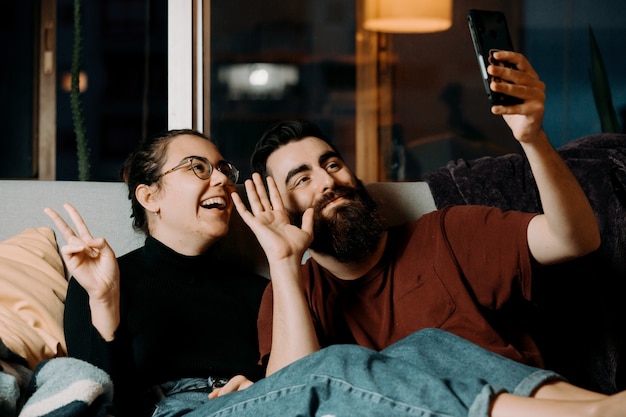 A young couple on the couch greeting to the mobile phone and smiling while on pandemic