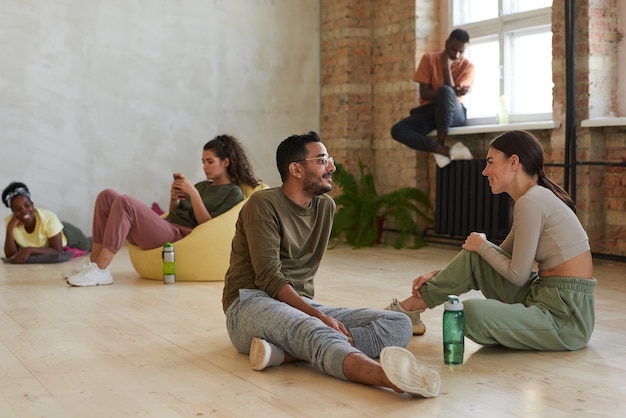 Young couple of dancers sitting on the floor and talking to each other in dance studio with other dancers 