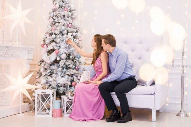 若いカップルがクリスマスツリーを飾ります。クリスマスの部屋の男性と女性のクリスマスのための派手な服。