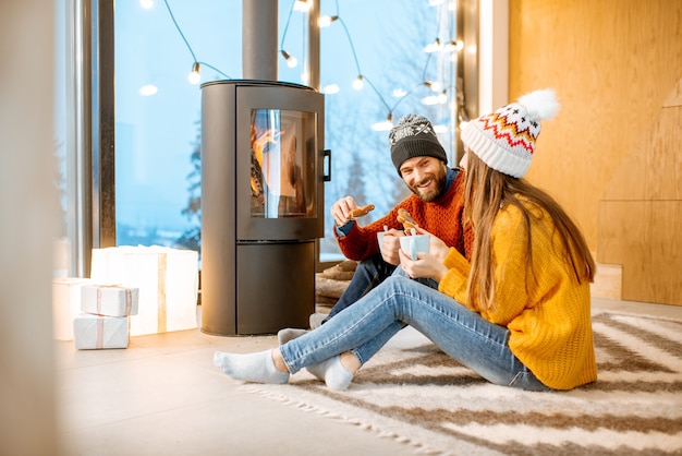 Young couple dressed in bright sweaters and hats sitting together near the fireplace in the modern house durnig winter time