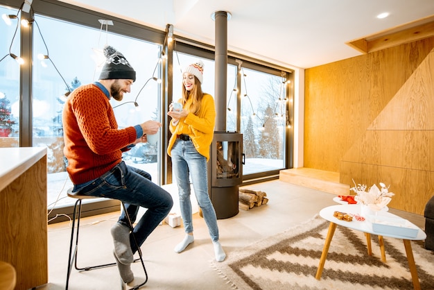 Young couple dressed in bright sweaters and hats standing together with hot drinks near the fireplace in the modern house durnig winter time