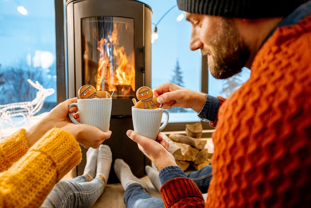 写真 冬の間に山のモダンな家の暖炉のそばでホットドリンクと明るいセーターを着た若いカップル
