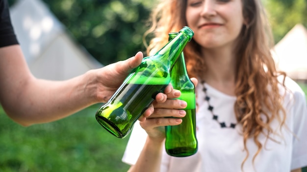 Young couple drinking beer, smiling, clinking bottles. greenery around. glamping