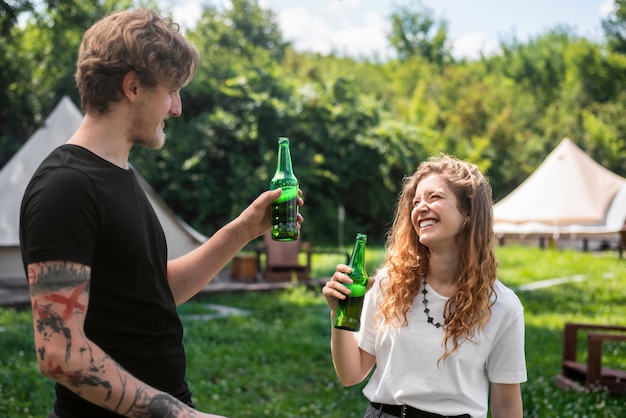 Young couple drinking beer, smiling. Greenery around. Glamping