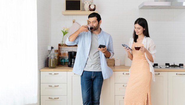 Young couple drinking hot coffee in the kitchen with smartphone Latin woman smiling looking at boyfriend while sitting on kitchen counter