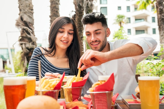 Young couple eating crispy French Fries or potato chips at an outdoor table at a restaurant or pub in a city accompanied by glasses of cold beer